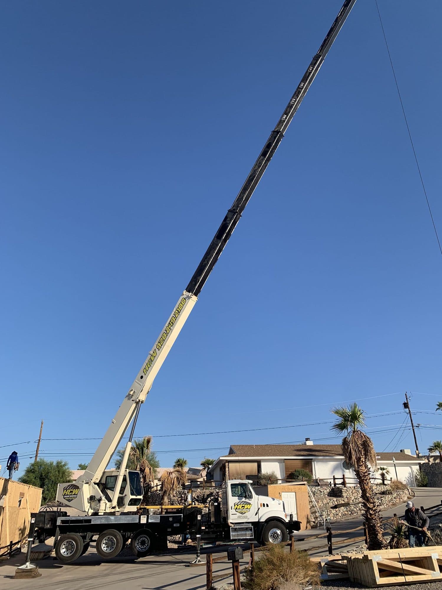 Boom truck crane with its boom fully extended into a clear sky above a residential framing site