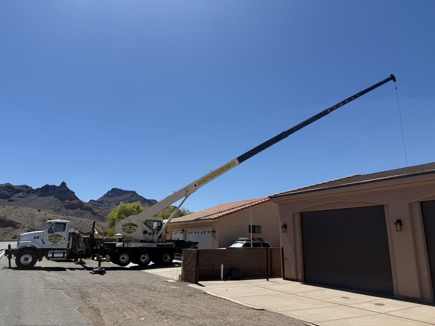 Boom truck crane reaching its long boom over a stucco garage with desert peaks in the background