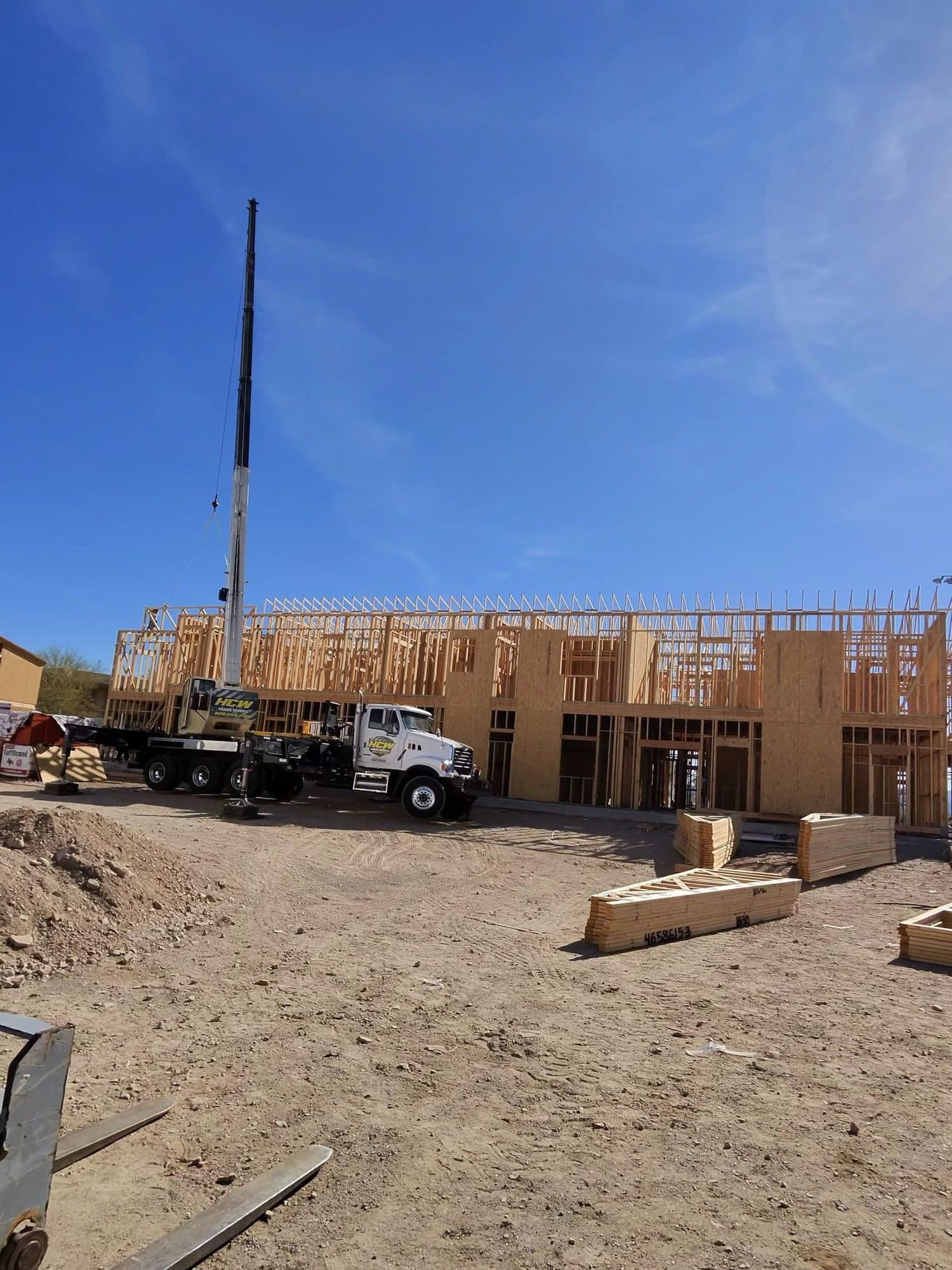 White HCW boom truck with boom raised parked before a large wood framed building with truss bundles on dirt