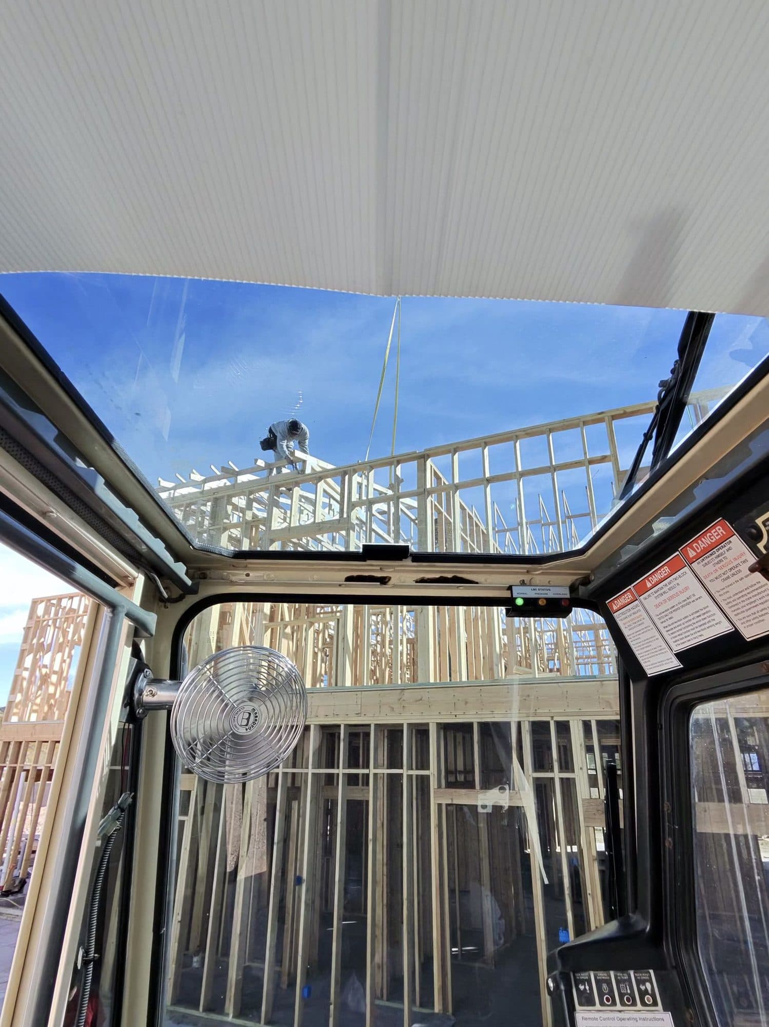 Through the crane cab roof window a framer works on a tall wood wall frame beside the hoist line