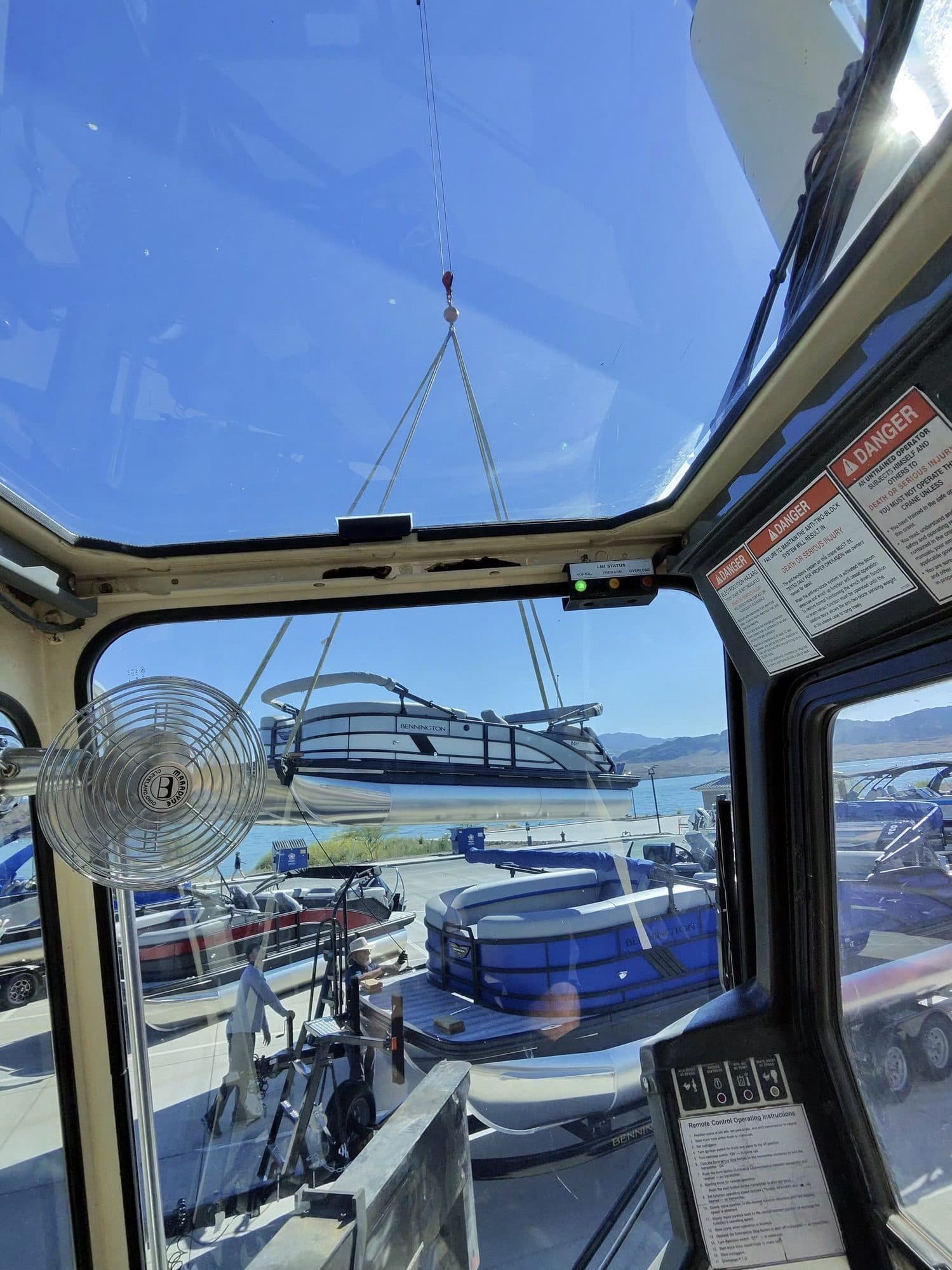 Pontoon boat hanging from crane slings above a lakeside lot of boats, seen from inside the crane cab