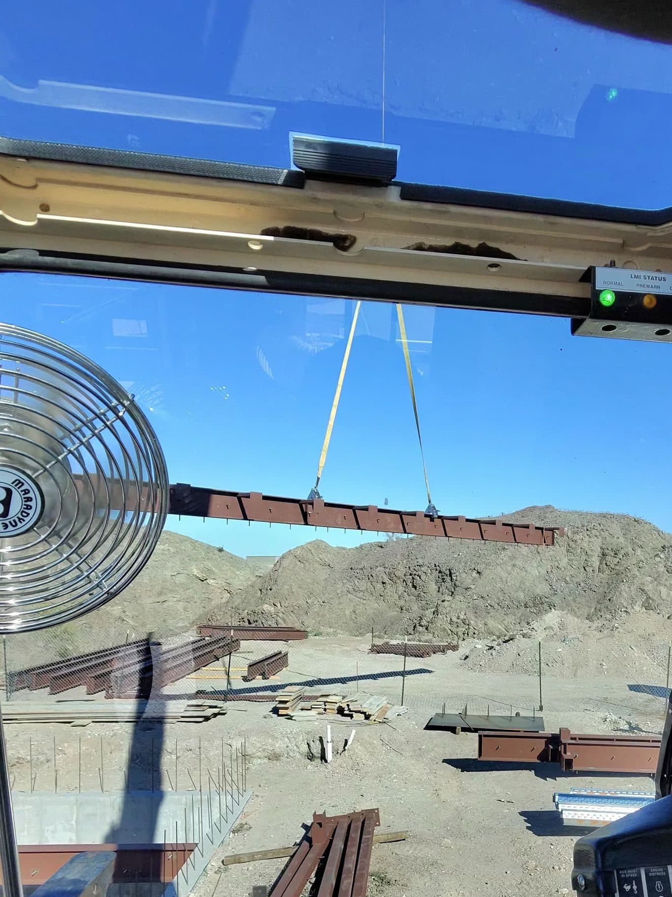 Steel beam hanging from yellow slings seen through the crane cab window, with the operator fan and controls