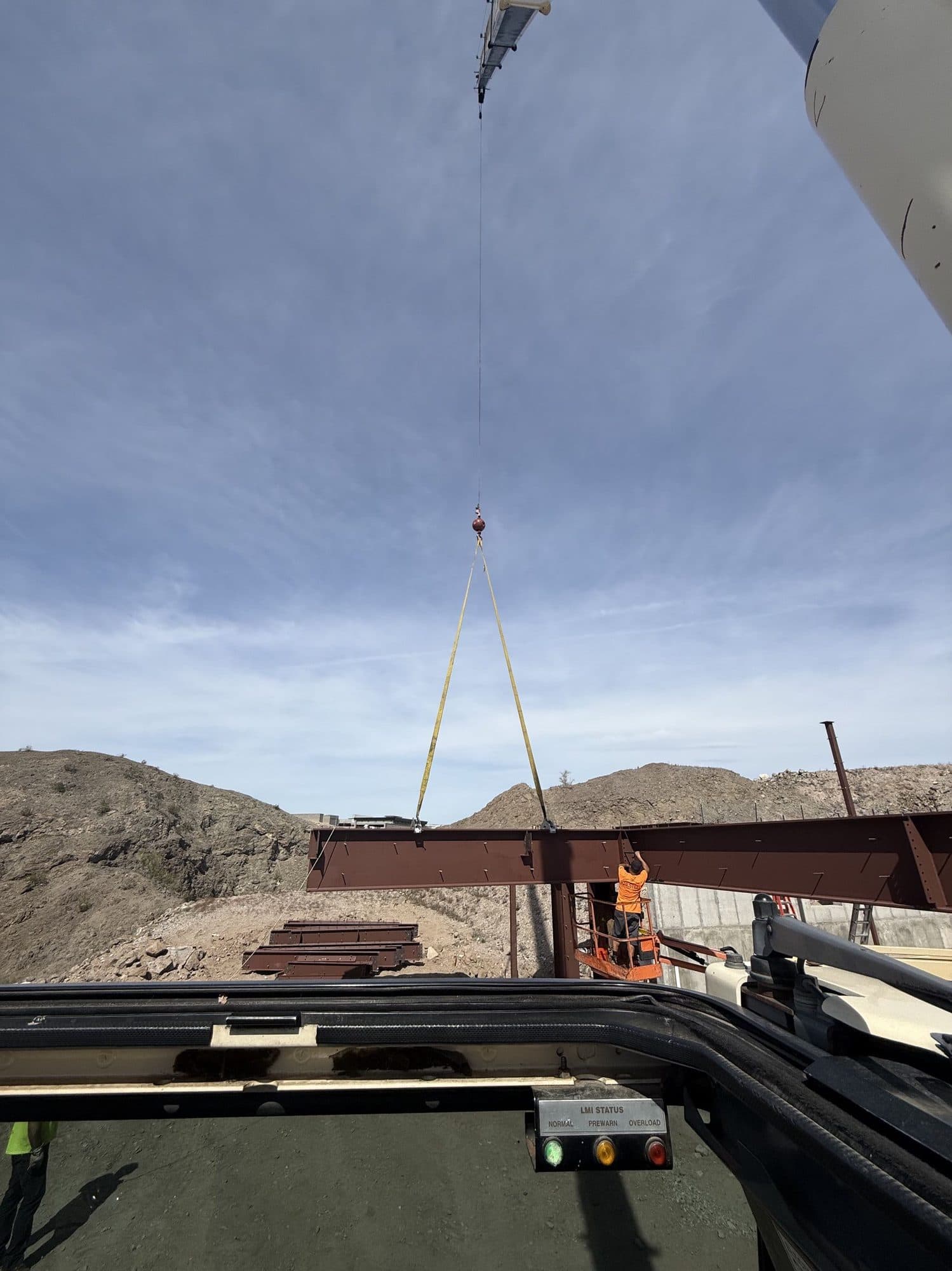 From the crane cab, a steel beam hangs from slings as a crew member on a lift guides it