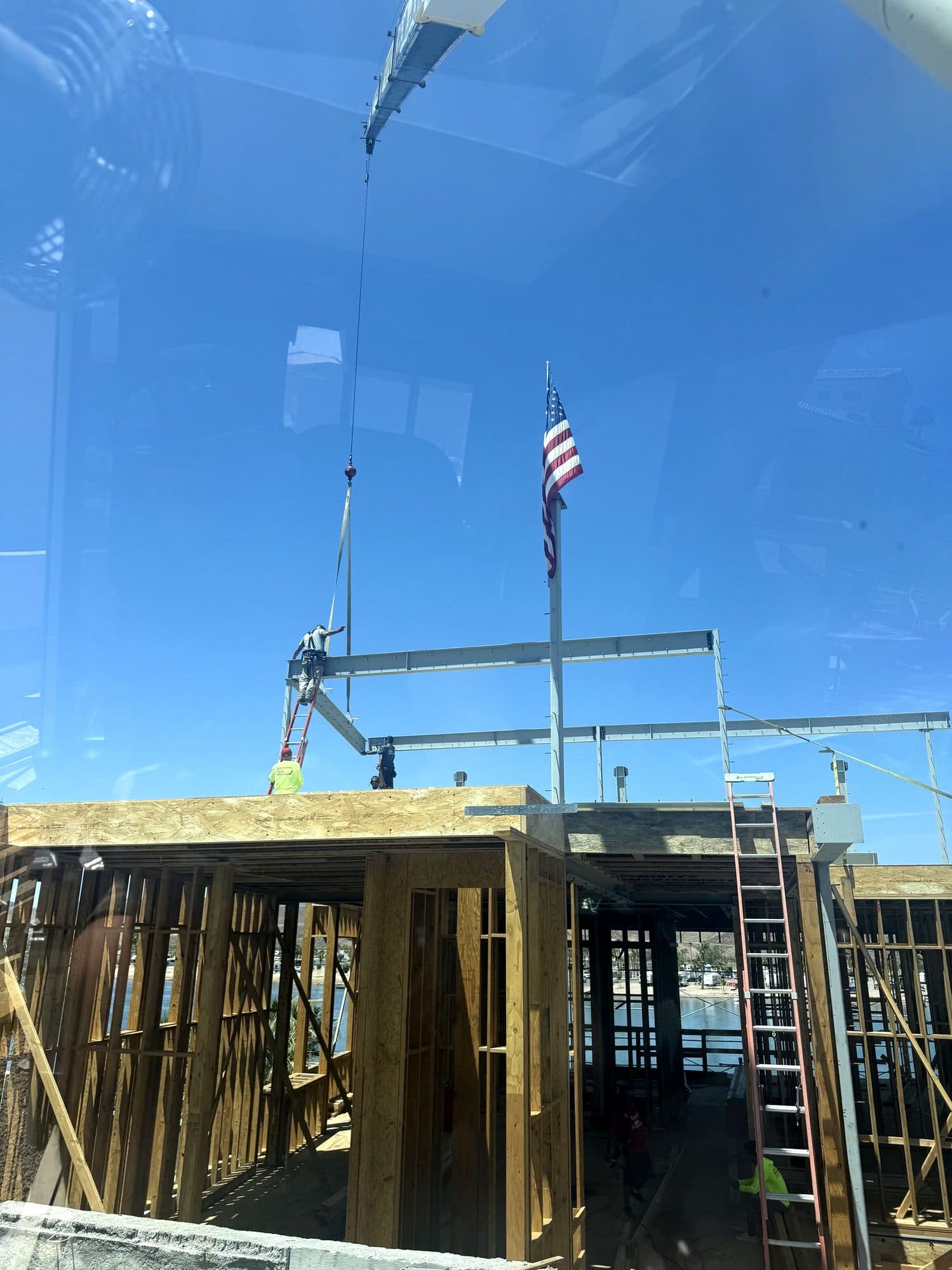 Through the crane cab window, crew set steel beams on a wood framed building near an American flag