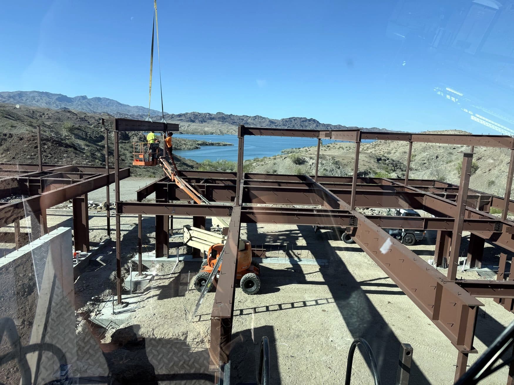 Steel building frame seen through the crane cab glass, crew on an aerial lift and a lake beyond