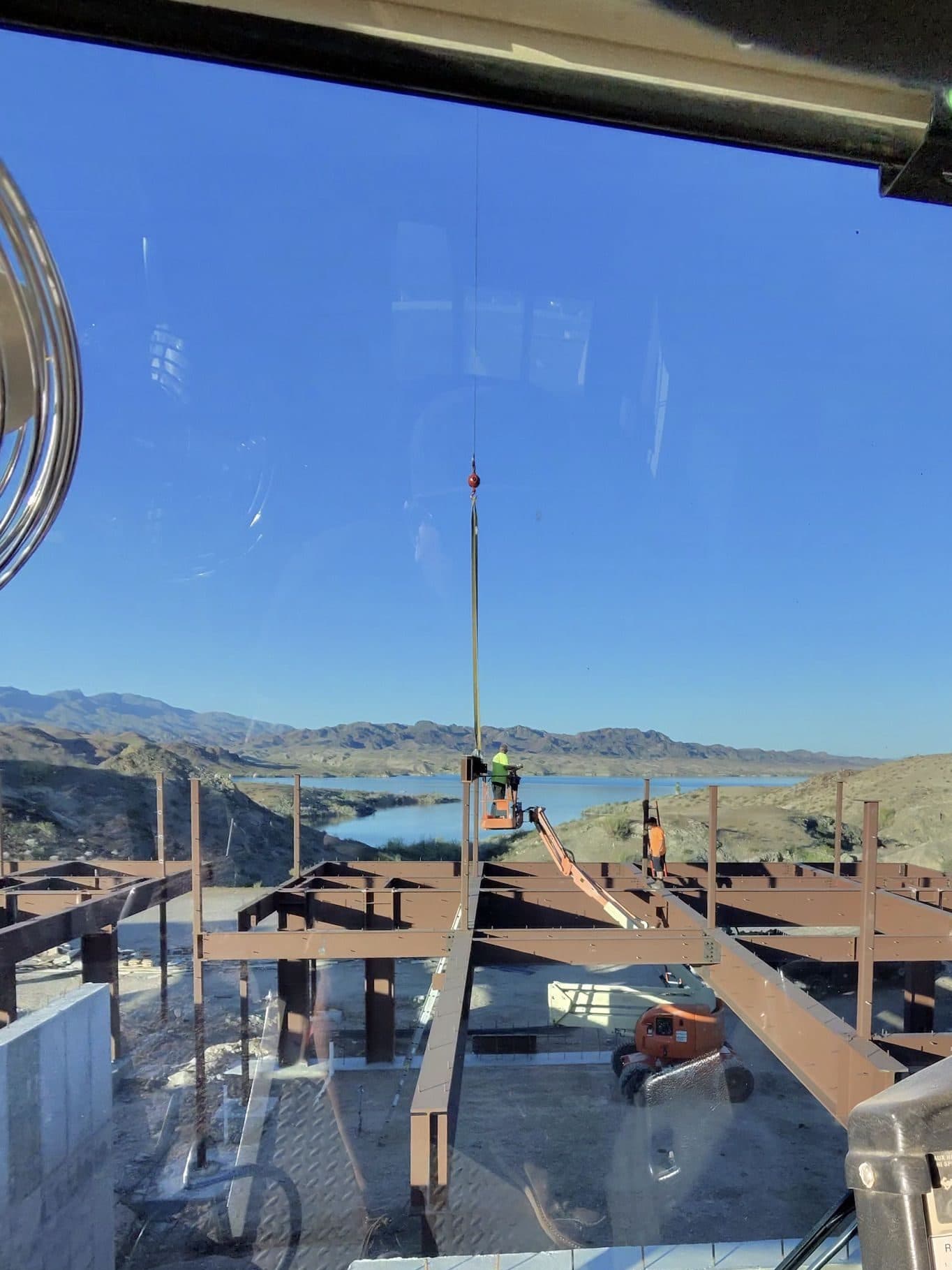 From the crane cab, a worker in a lift guides the hook over a steel frame beside the lake