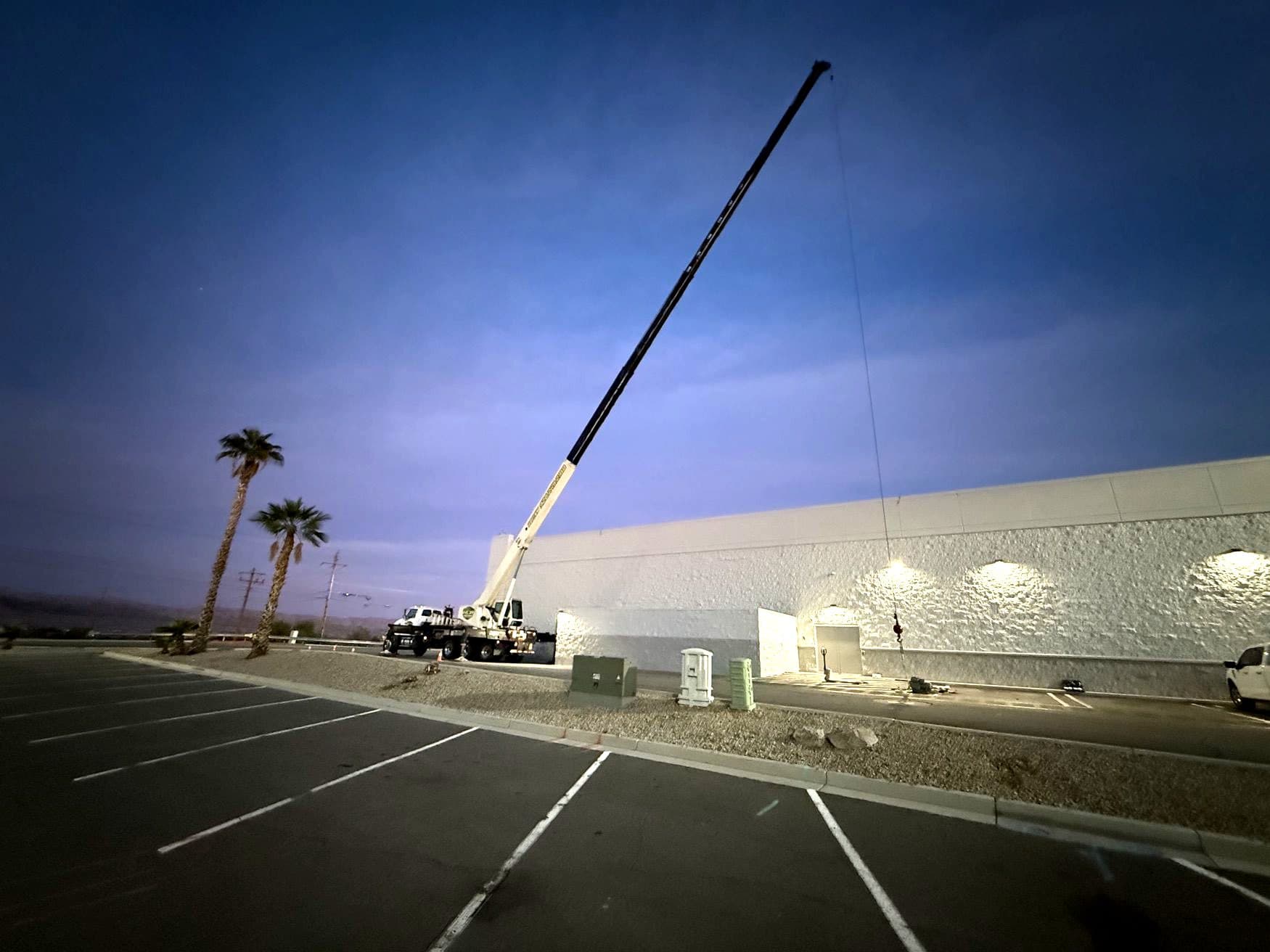 Mobile crane with long extended boom beside a large commercial building at dusk, seen across a parking lot