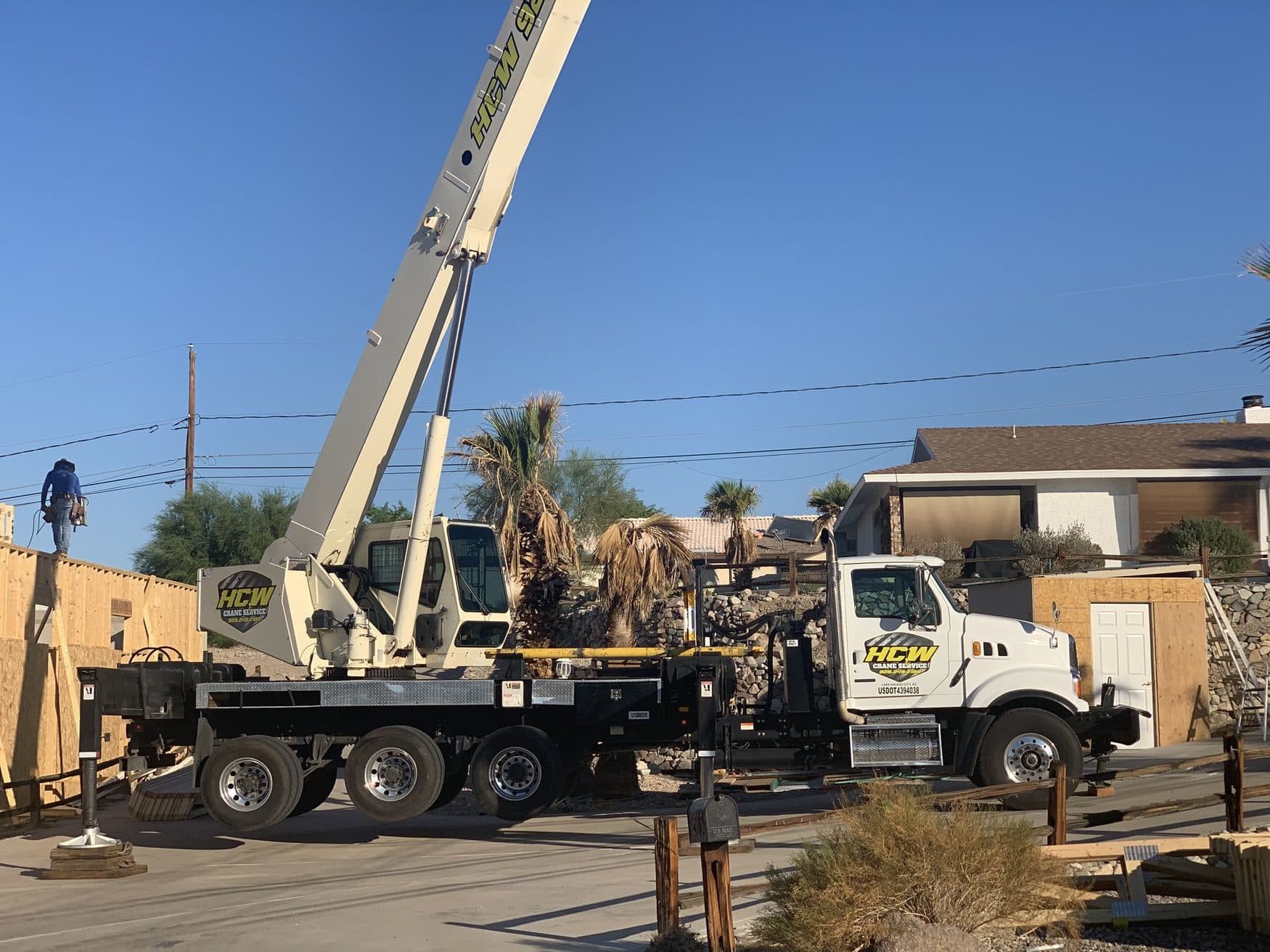White boom truck crane set up on a residential street beside wood wall framing with a worker on top