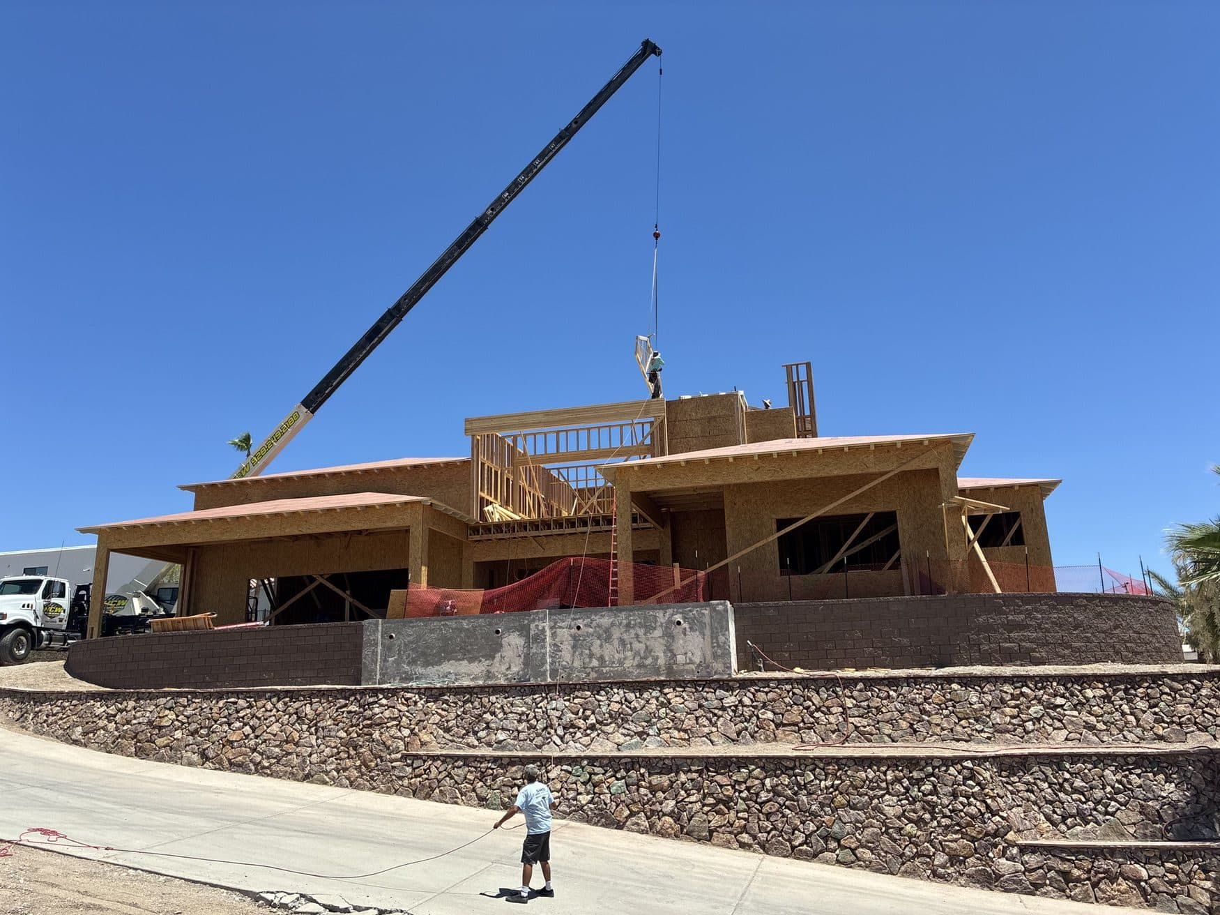 Crane boom lowering framing material to workers on a hillside home, ground crew guiding with a tag line