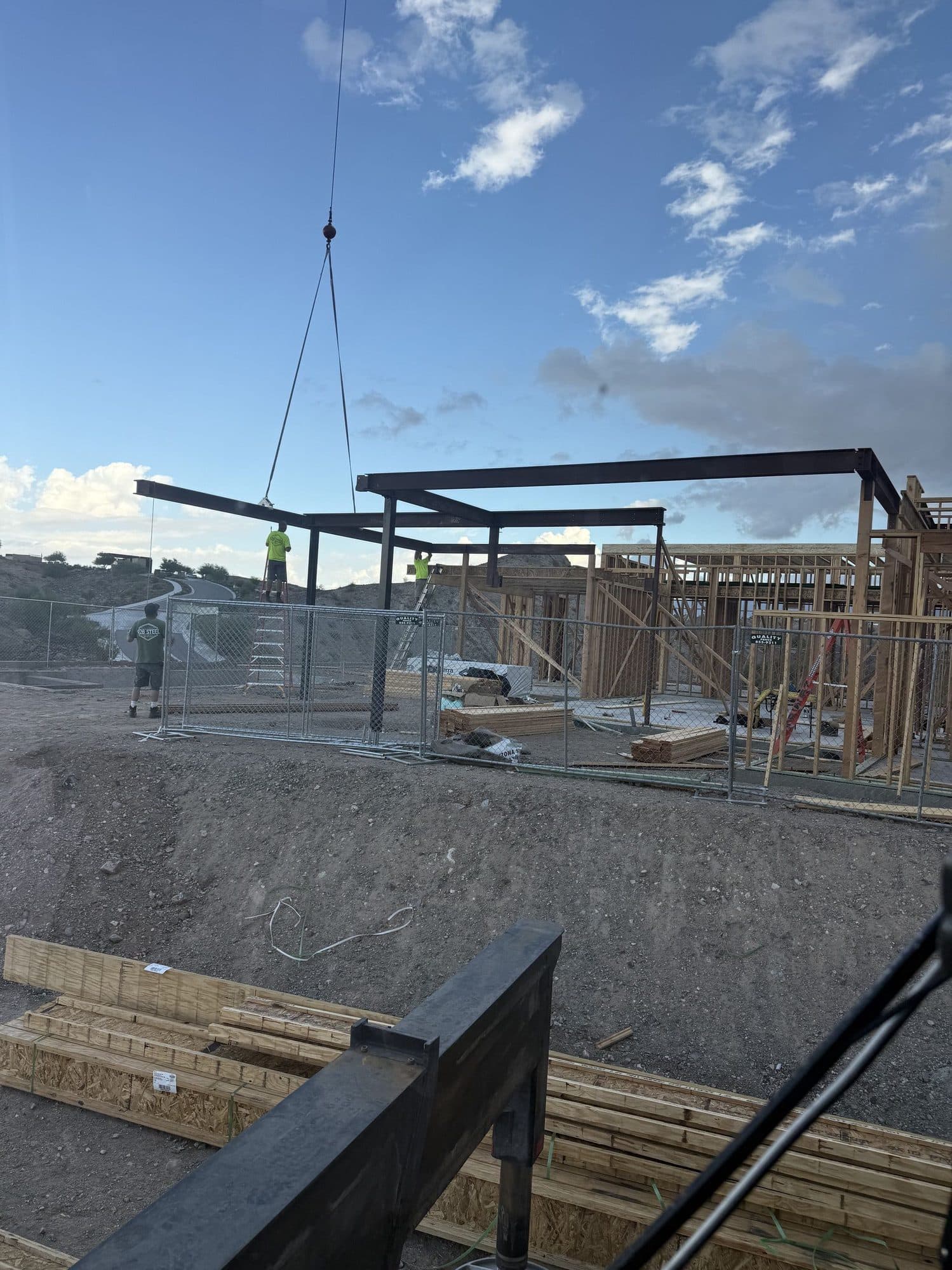 Crane hook and slings lowering a steel beam onto a hillside steel frame while crew on ladders guide it