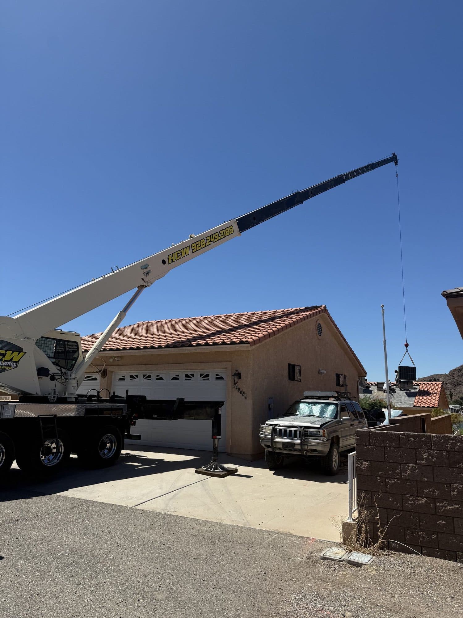 Boom truck crane on a driveway lowering an HVAC unit over a tile roof garage to a neighboring roof
