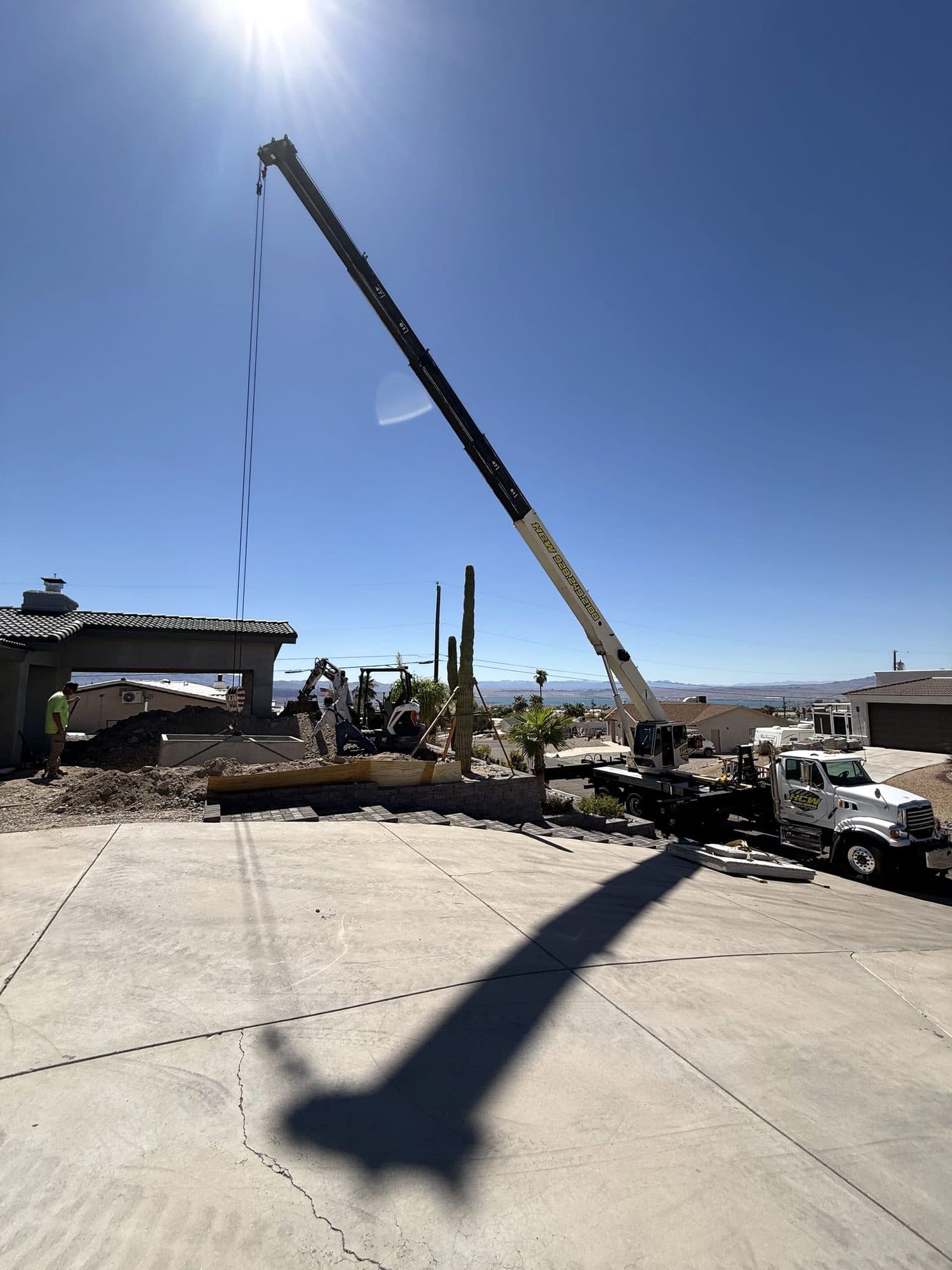 Crane boom lowering a concrete piece into a hillside landscape project with an excavator and crew nearby