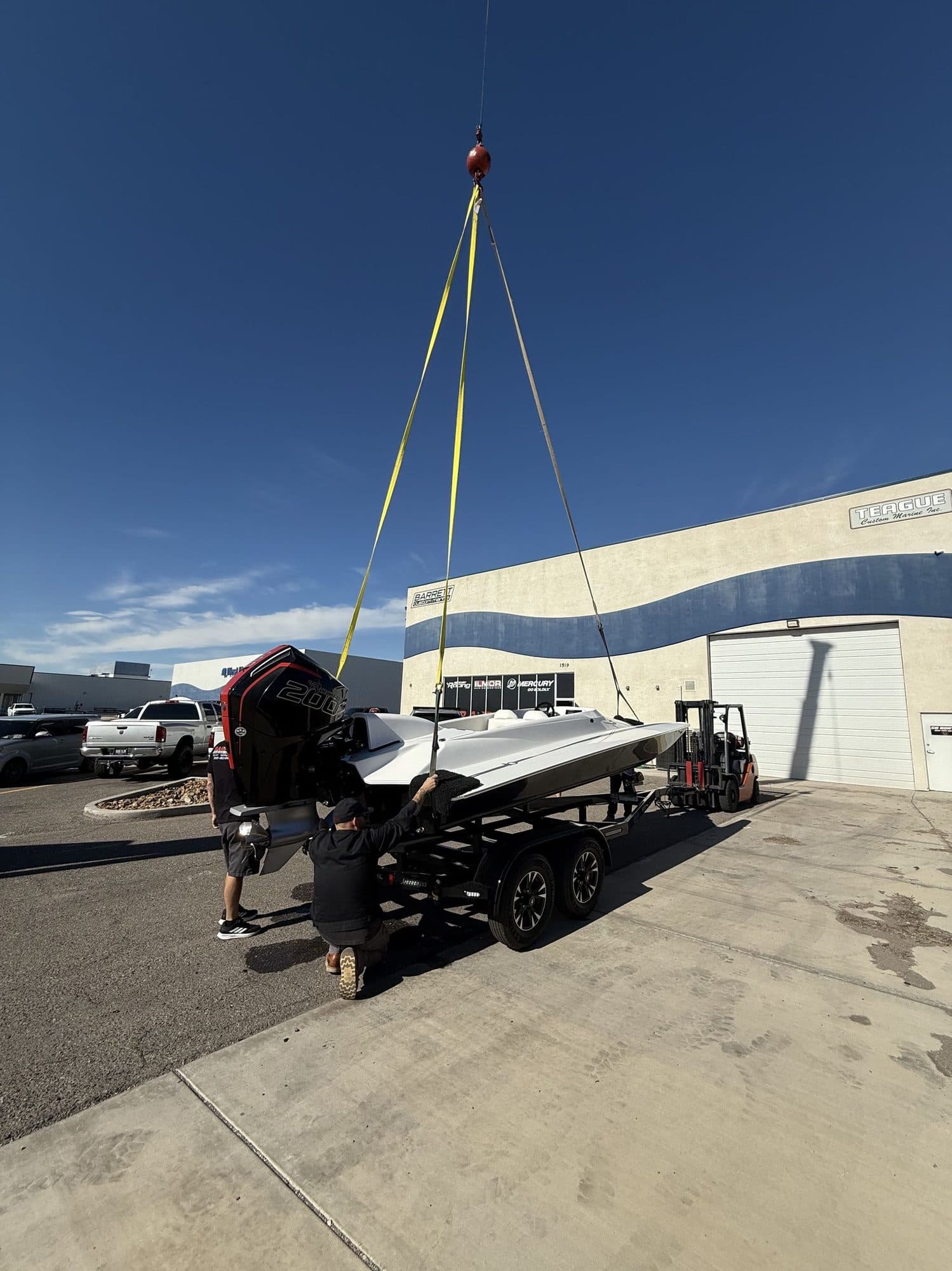Yellow lifting slings from a crane hook raise a white race boat off its trailer while two crew steady it