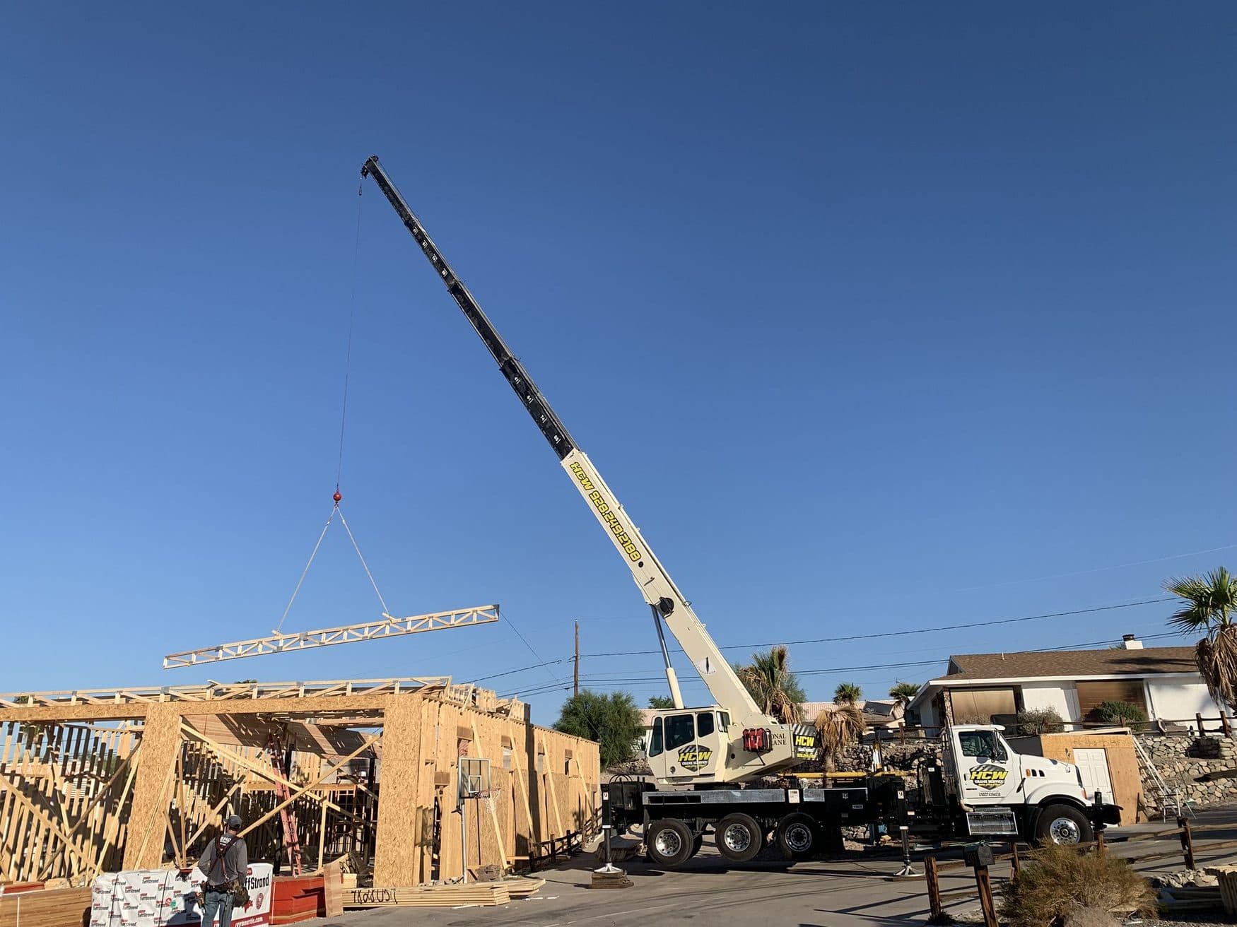 Boom truck crane lifting a wood roof truss over freshly framed walls while a worker watches below