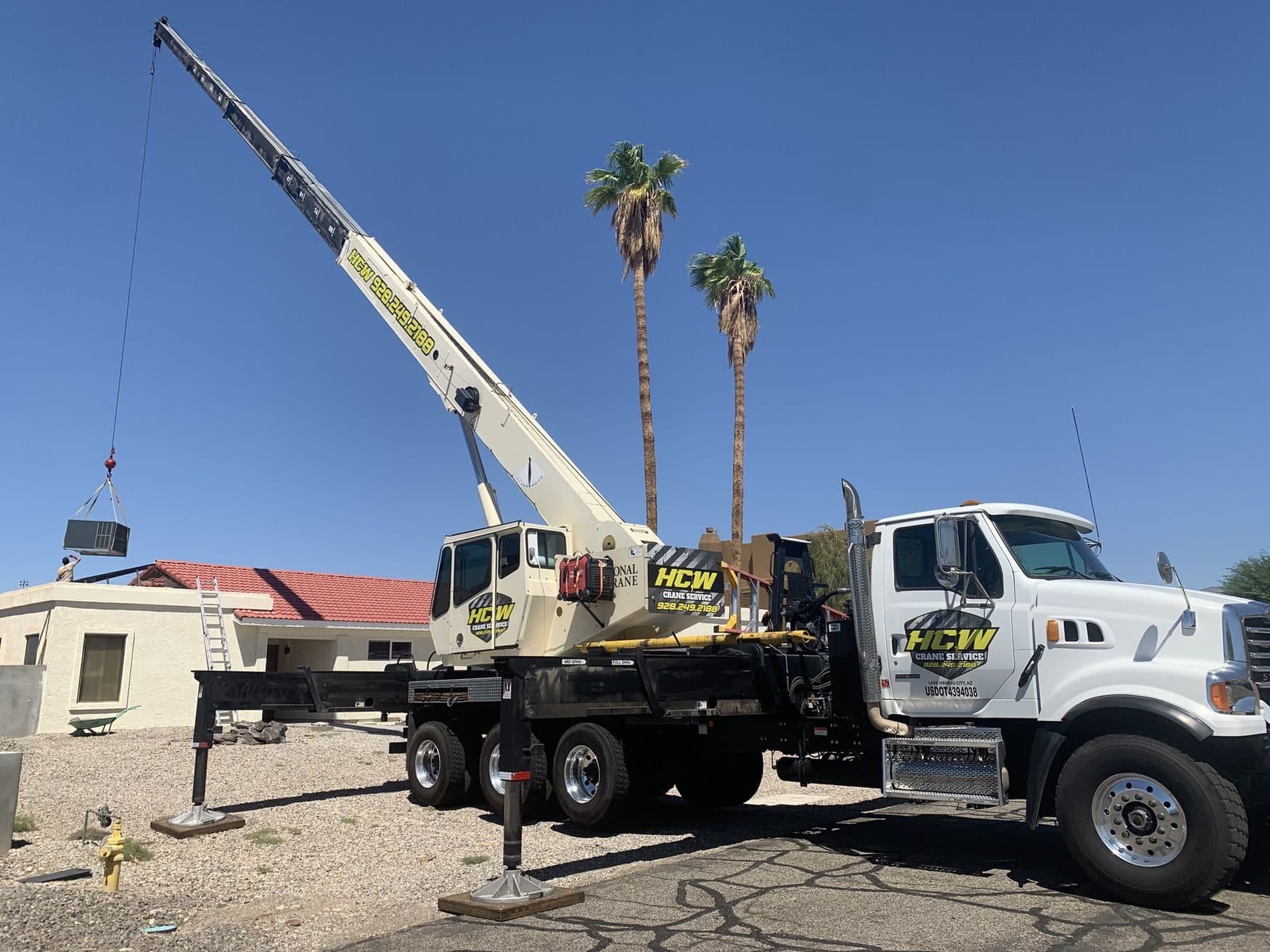 HCW boom truck crane on outriggers and cribbing swinging an HVAC unit over a red tile roof