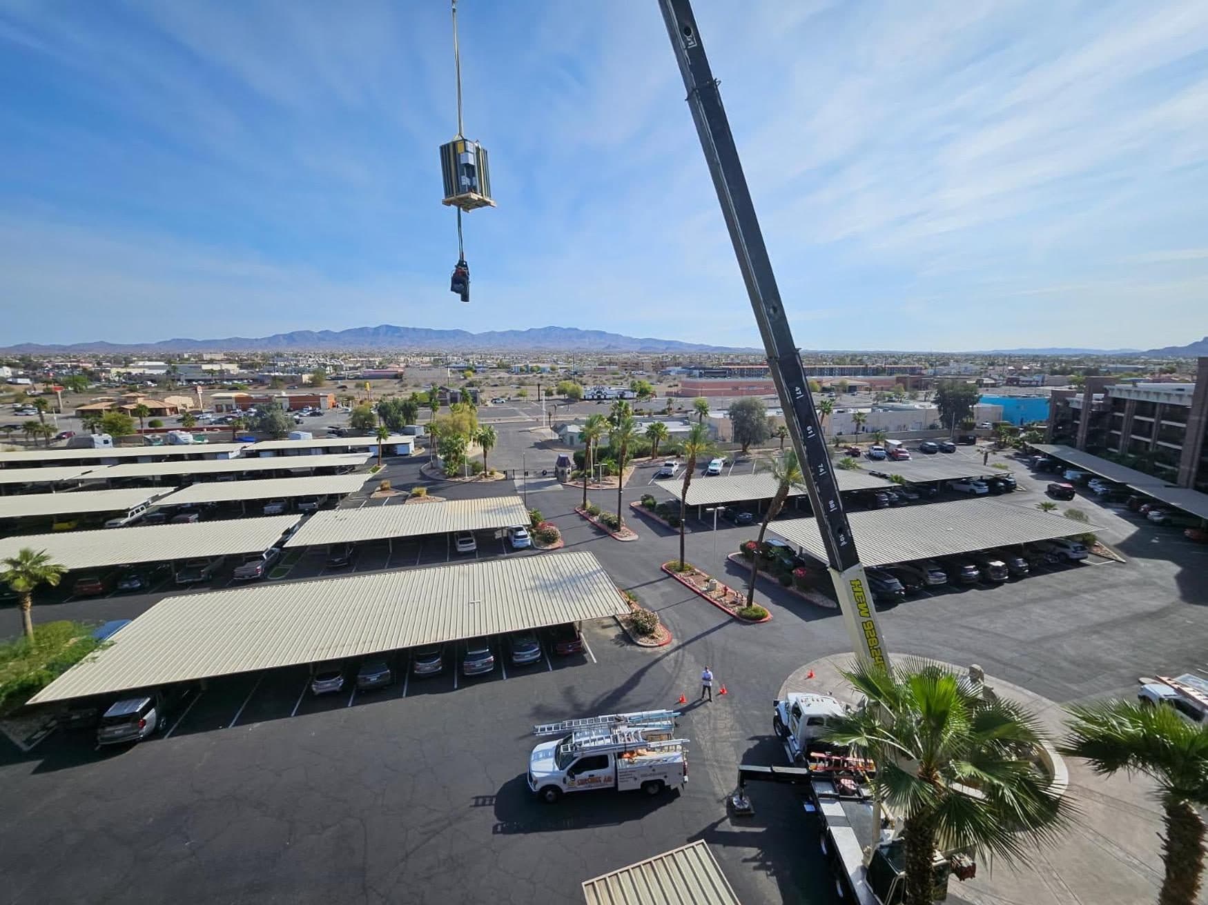 Rooftop HVAC unit on a pallet hanging from the crane line high above a commercial parking lot, boom truck below