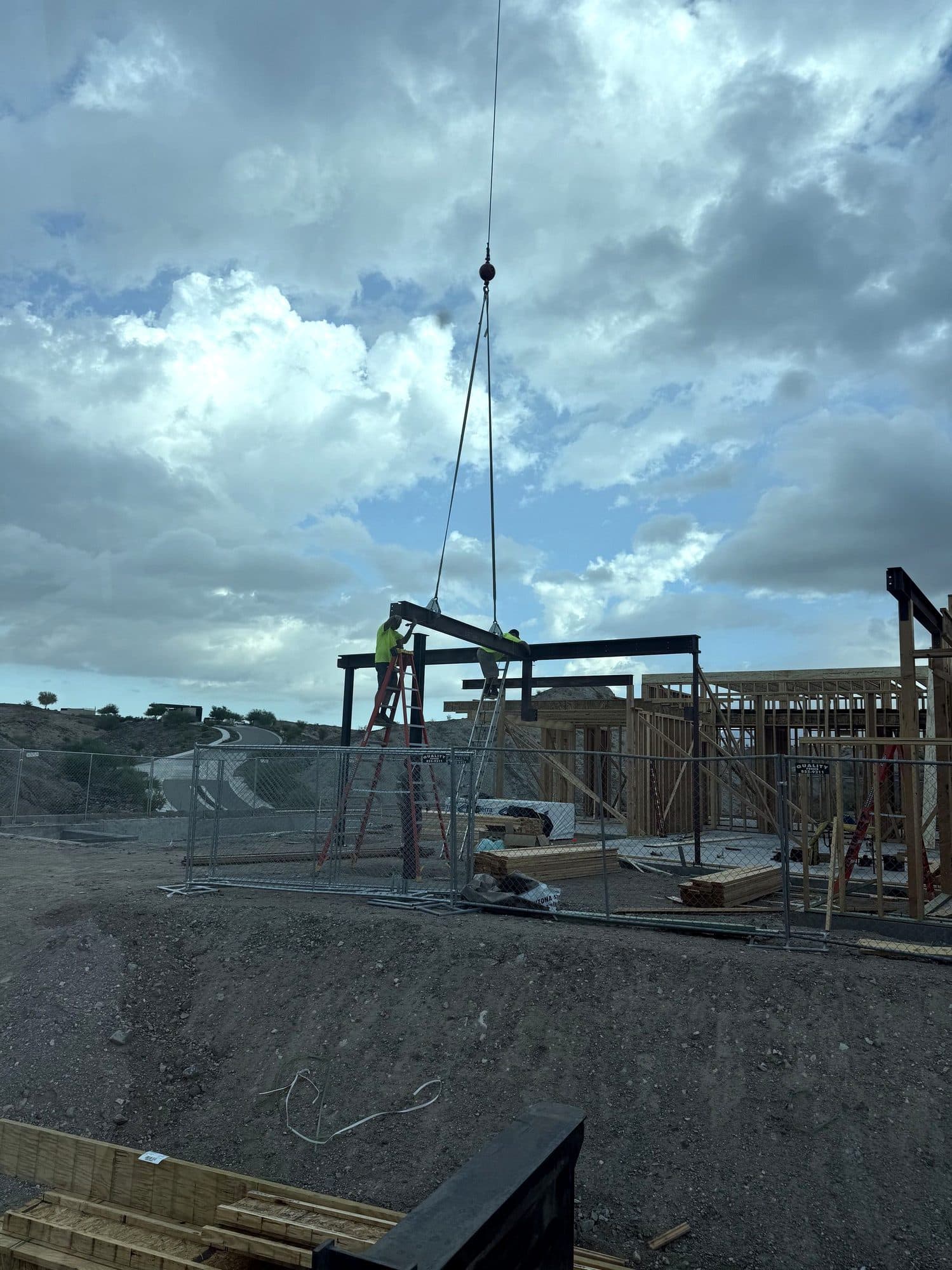 Crane rigging lowering a steel beam to two workers on ladders at a hillside steel frame