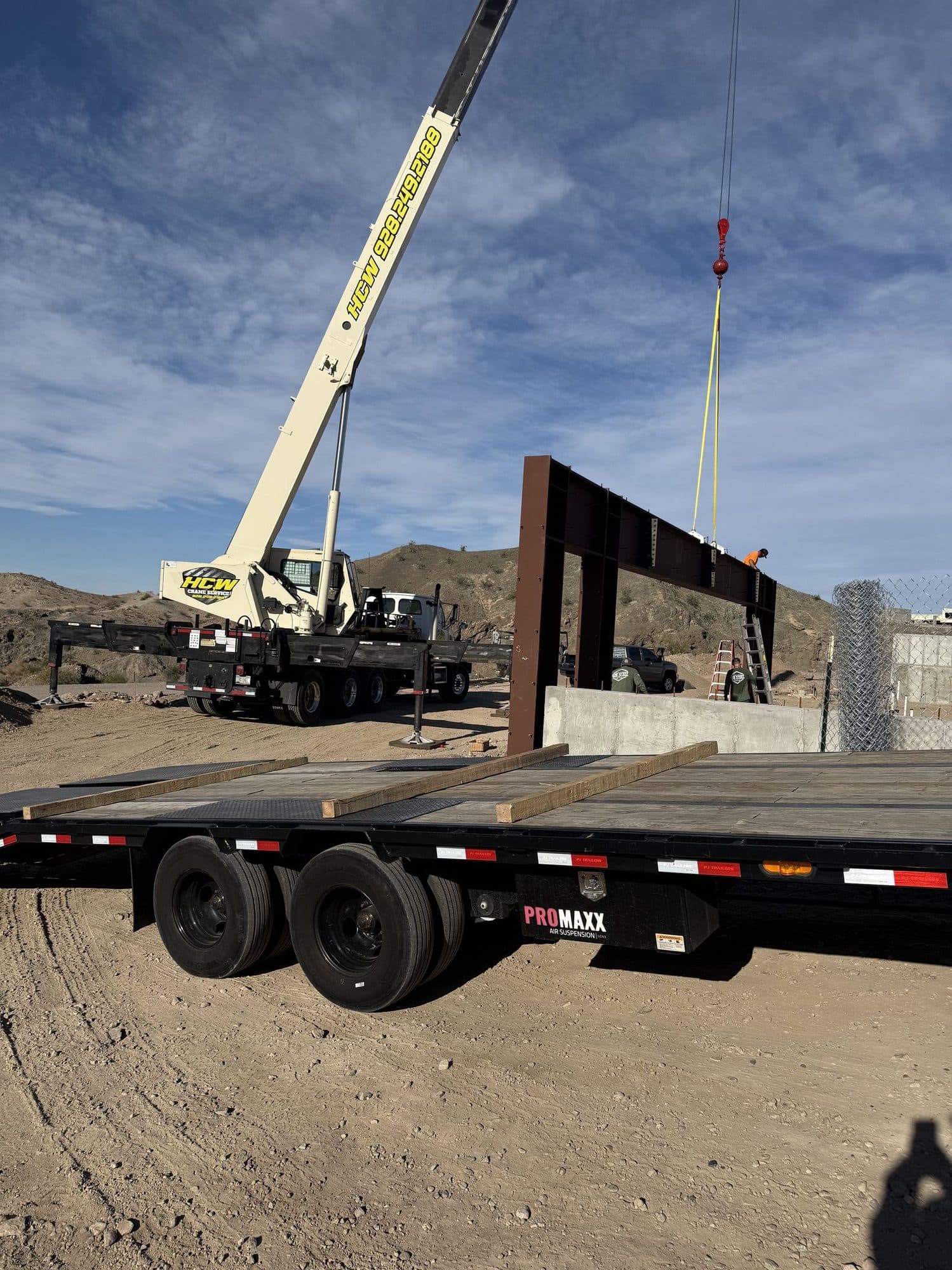Boom truck crane setting a steel frame on a foundation, with an empty flatbed trailer in the foreground
