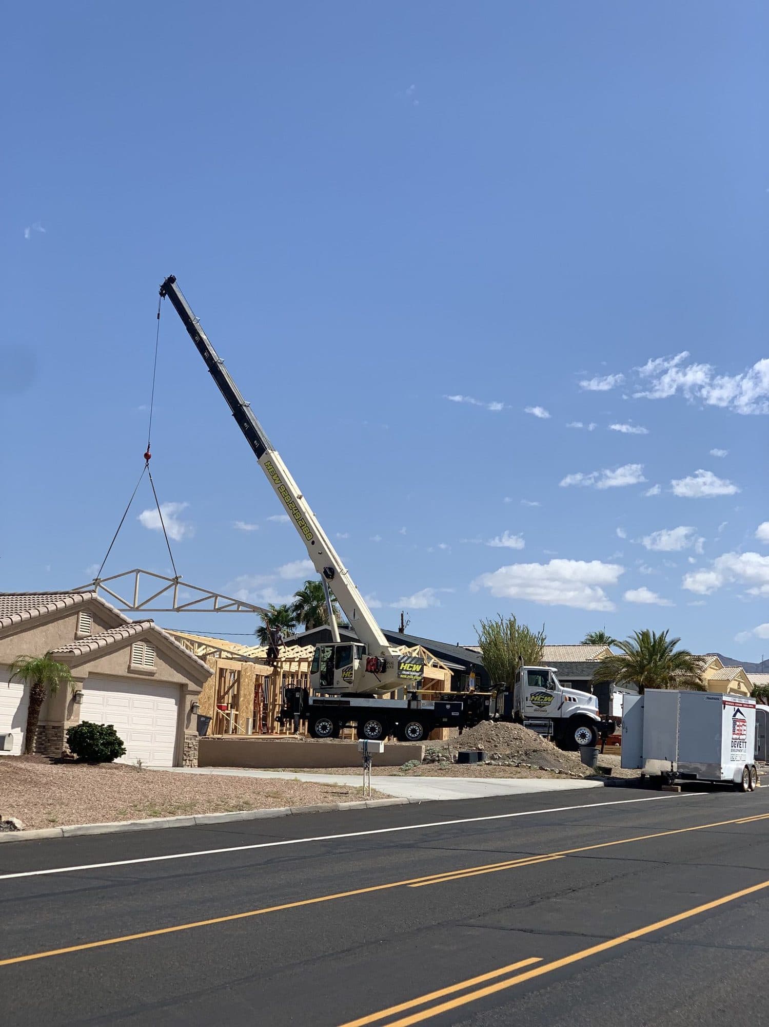 Boom truck crane lifting a roof truss over a house onto new framing beside a neighborhood street