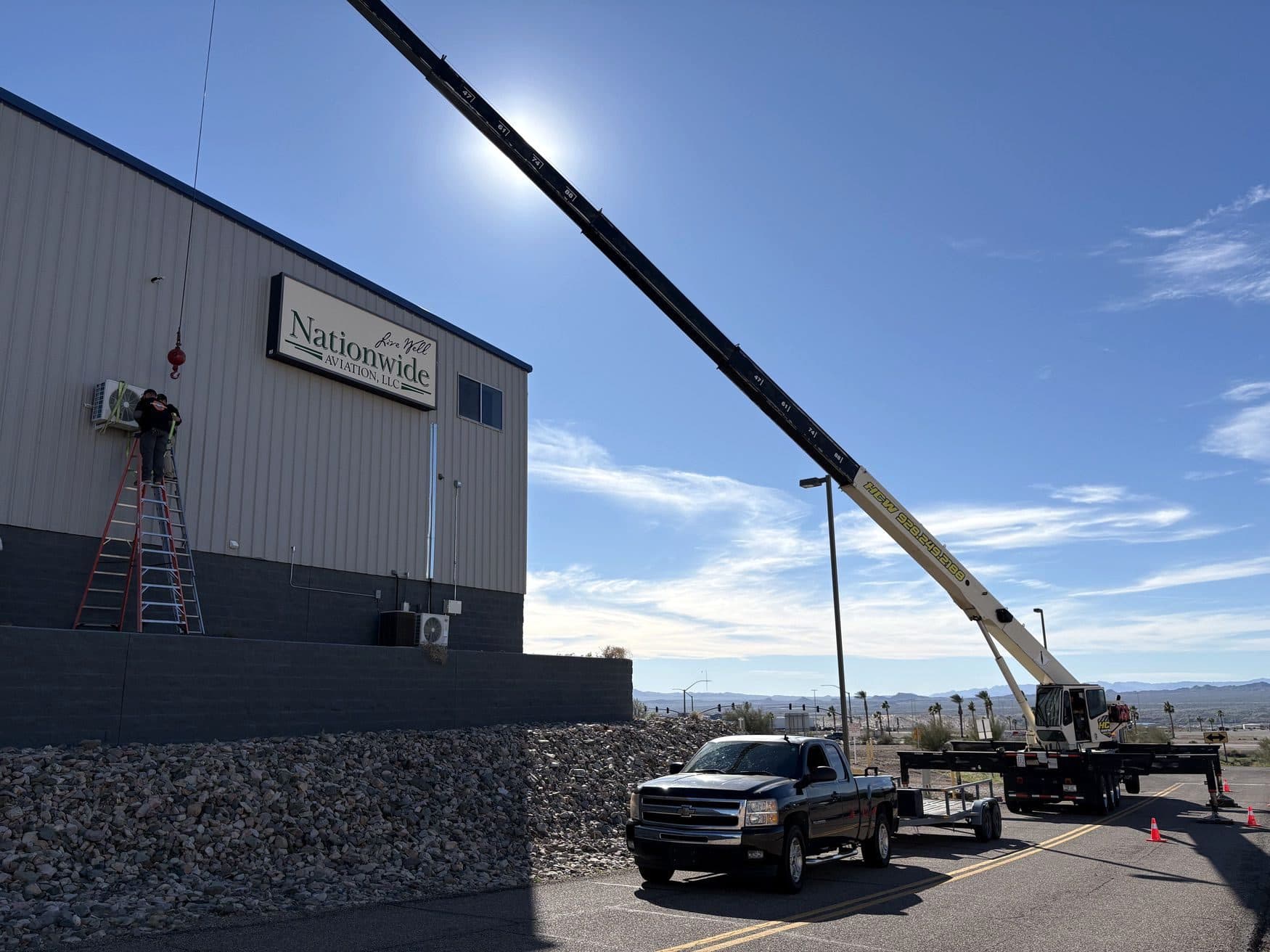 Crane hook beside a worker on a ladder placing a wall mounted condenser, boom truck parked down the road
