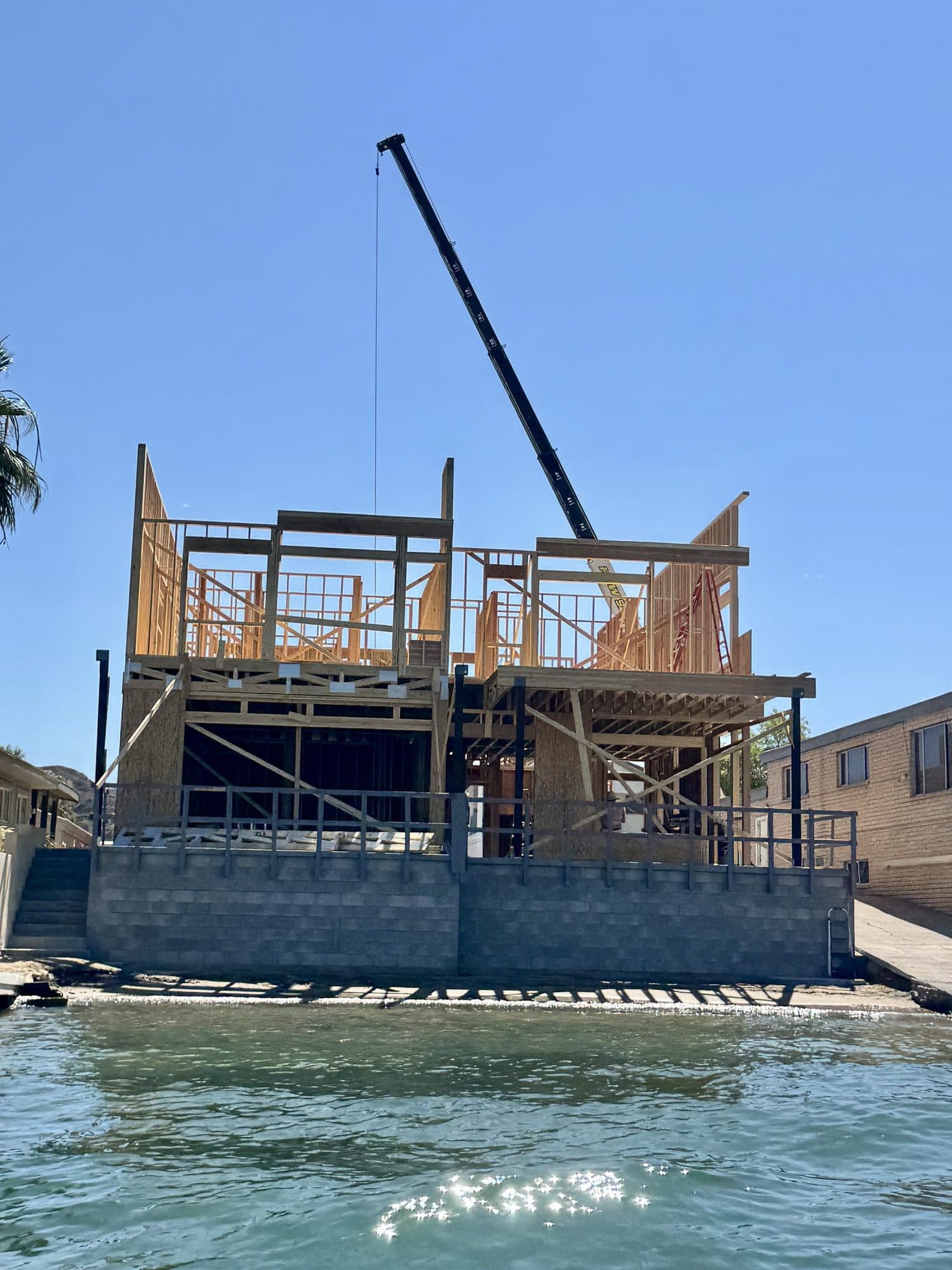 Crane boom rising behind a two story wood framed waterfront home under construction, seen from the water