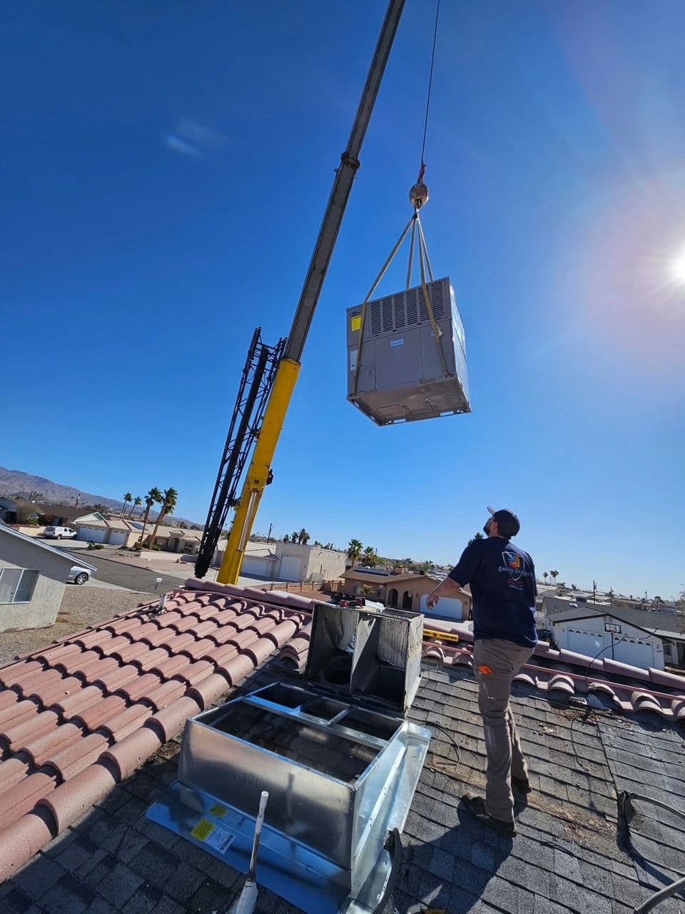 Crane lowering a rooftop package AC unit to an open roof curb as a technician guides it