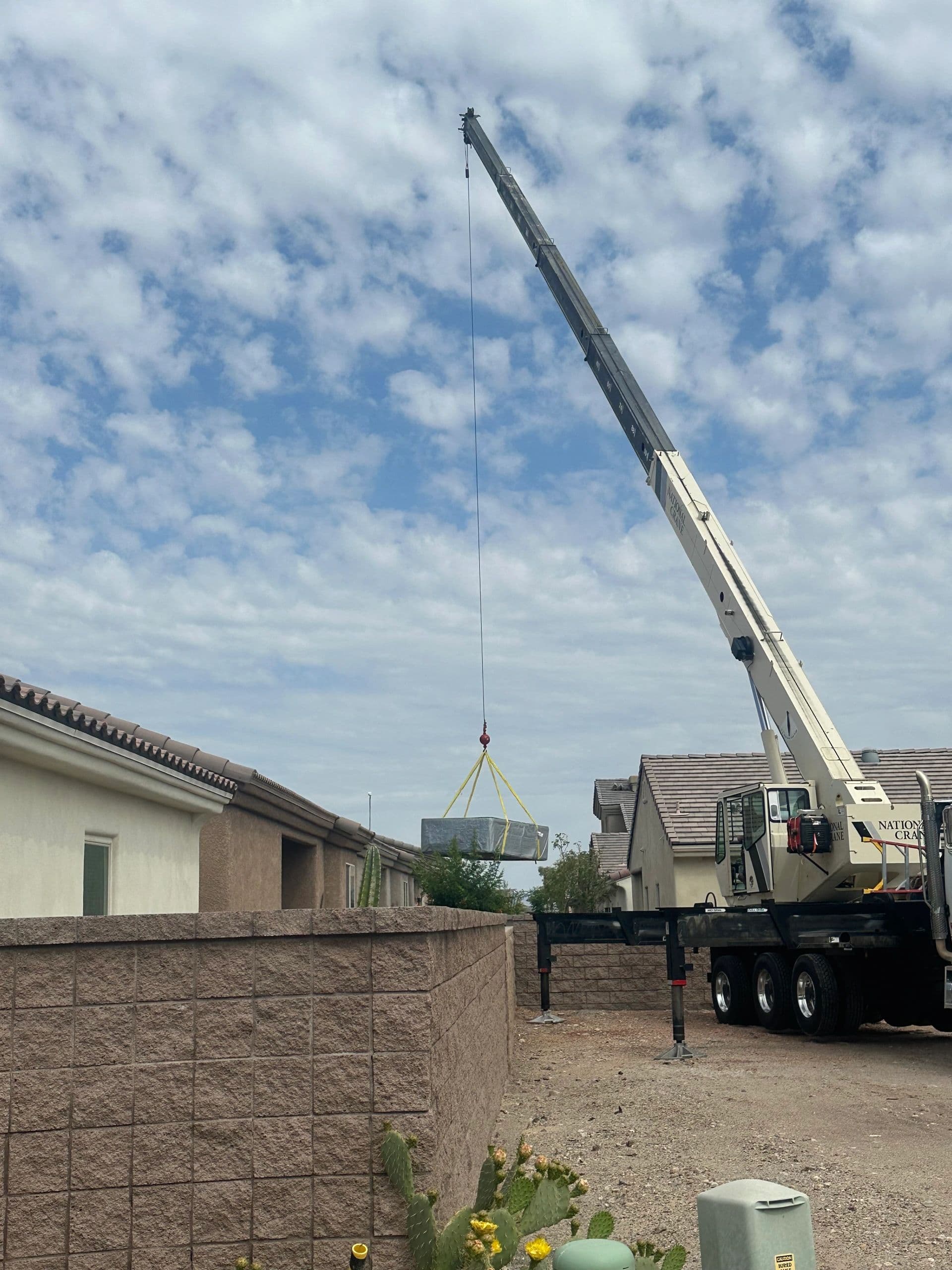 Crane lowering a wrapped load over a block wall into a backyard between two homes