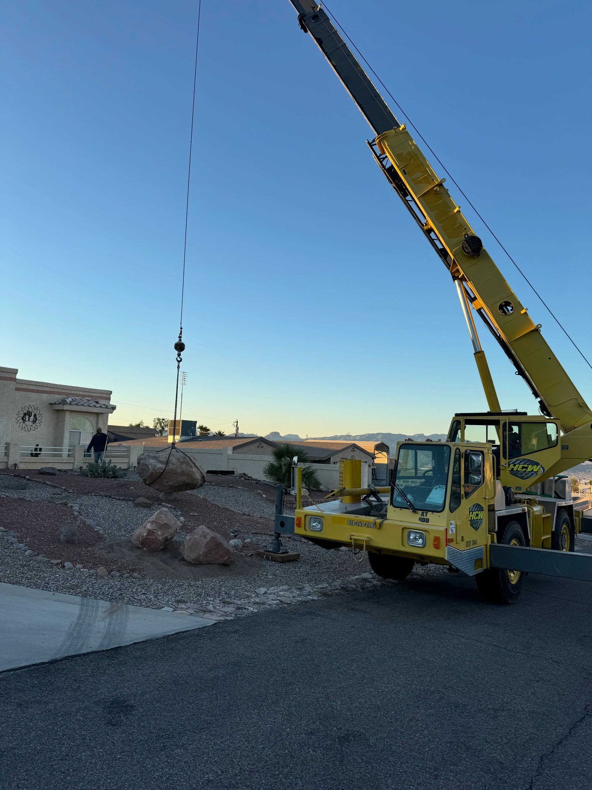 Rough-terrain crane setting a large landscape boulder in a desert front yard at dusk