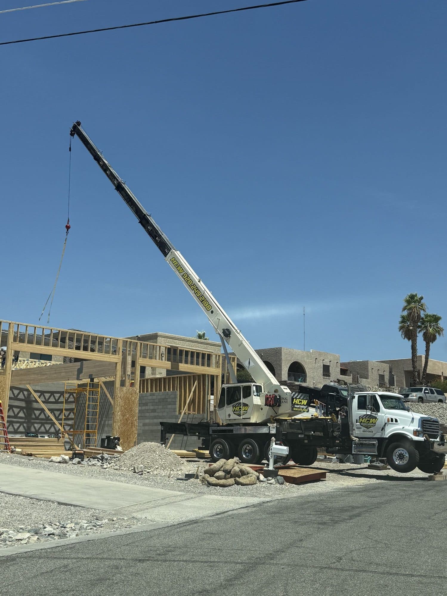 HCW boom truck crane on outriggers with boom extended over a wood framed home under construction