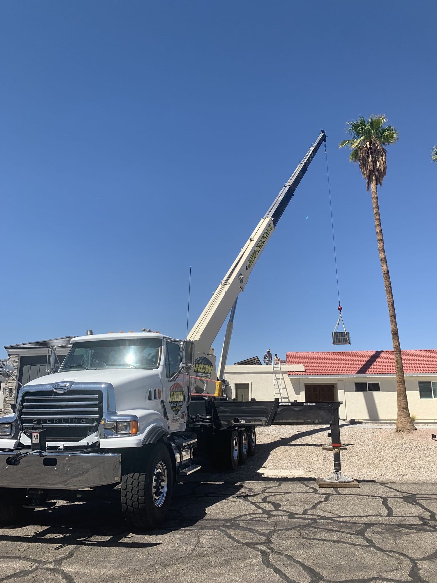 White boom truck crane lifting a rooftop HVAC unit toward a red tile roof beside a tall palm