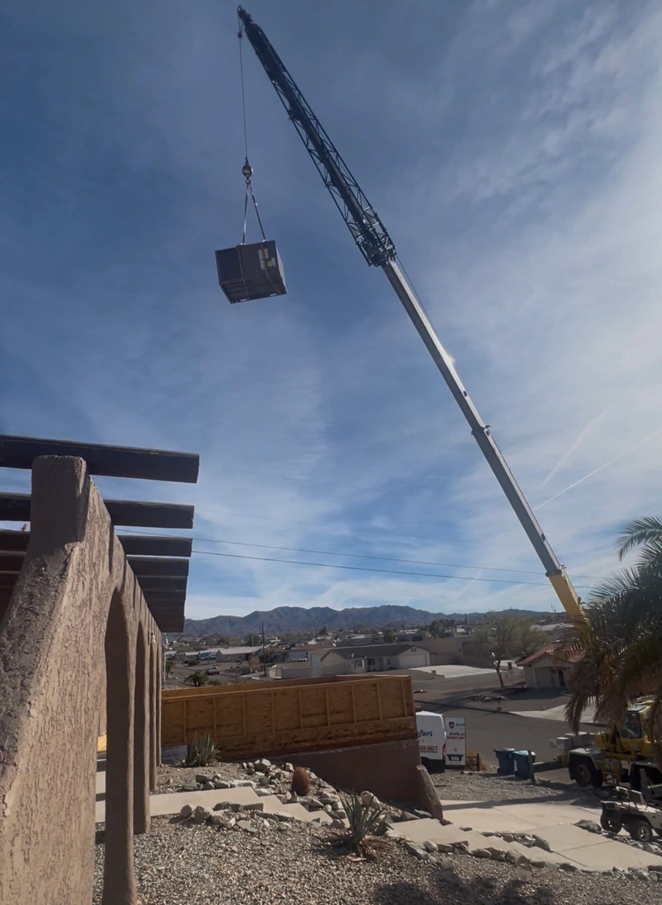 Crane hoisting an HVAC unit high above a desert neighborhood under blue sky